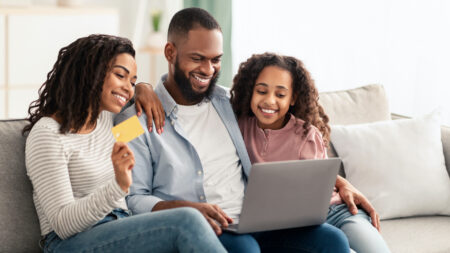 Easy Payment And Cashback Concept. Portrait of smiling African American family doing online shopping at home using laptop, woman holding credit card in hand, people sitting on couch at home