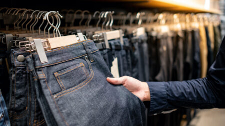 Male hand customer choosing black men jeans or denim pants (trousers) hanging on rack in clothes shop. Fashion product collection in clothing store for selling. Textile industry and business concept